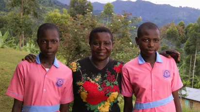 Portrait de Rebecca Nakayega Wandegge, dans une zone rurale, souriante, à côté de ses deux filles qui portent leur uniforme scolaire. (Ouganda, 2025)